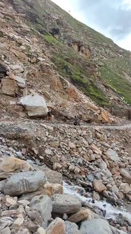 A person on a motorcycle rides along a narrow dirt road winding through a steep, rocky mountainside. The road is covered in loose rocks and debris, suggesting a recent landslide or rockfall. Large boulders are scattered on the road and down the slope. The mountainside is covered in patches of green vegetation. The sky is overcast. The motorcycle rider is wearing a helmet and a backpack. The rider appears to be navigating the treacherous terrain carefully.