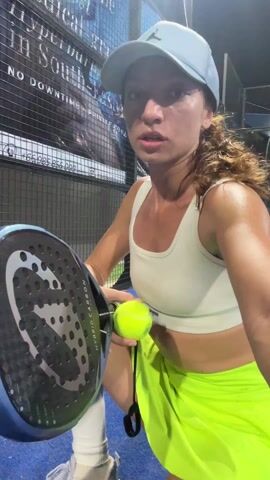 A woman is on a blue tennis court with black netting and a sign in the background. She is holding a tennis racket and a tennis ball. She bounces the ball on the racket, catches it in her mouth, then drops it and hits it back towards the camera. She smiles at the camera at the end. She wears a light blue hat, a white sports bra, a neon yellow skirt, and white socks and tennis shoes.