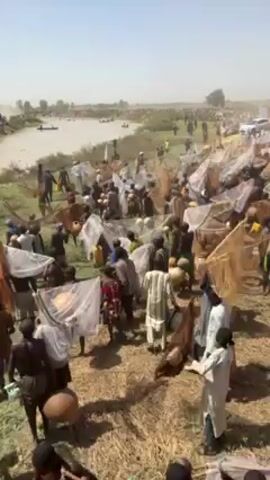The video shows a large crowd of people, mainly men, gathered on the bank of a river or large body of water. They are predominantly dark-skinned, and many are carrying or using fishing nets. Some individuals wear traditional clothing. There are small boats on the water, and a few vehicles visible in the background. The environment appears dry and dusty with sparse vegetation. It seems to be a community fishing event.