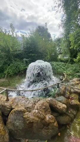 The video shows a close-up view of a fountain in a park or garden setting. The fountain is constructed from rocks and features a strong stream of water cascading down. The surrounding environment is lush and green, with trees and foliage visible in the background. The sky is cloudy. The camera appears to be positioned at ground level, looking up towards the fountain. There are no people visible in the video. The focus is on the natural elements of the scene, with the water flow being a prominent feature. The overall atmosphere is serene and naturalistic.