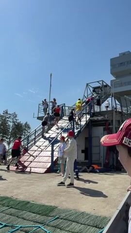 The video shows a group of people climbing a tall, metal structure with multiple staircases, resembling an outdoor obstacle course. Most of the climbers are dressed in athletic wear, some in shorts and others in long sleeves. There are observers on the ground near the base of the structure, some wearing hats. The sky is clear and blue, suggesting a sunny day. The structure is located in an outdoor area near a building.