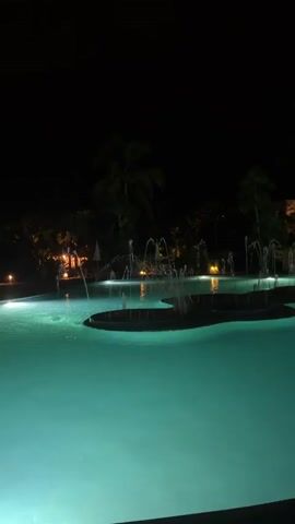 The video shows a dark scene of a swimming pool at night. The water in the pool appears turquoise under the artificial lighting.  Several small fountain jets are active in the middle of the pool. Palm trees and other foliage surround the pool area, illuminated by lights, while the sky is dark.