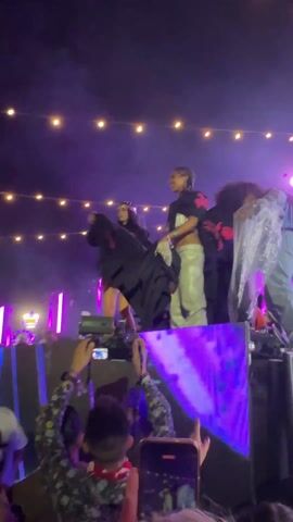 Several people stand on a platform, illuminated by bright, pulsating lights and string lights overhead. The crowd below raises their hands and phones to capture the scene. The people on the platform are dressed in trendy clothing, including silver pants and hooded tops. One person removes a garment and throws it into the crowd. The scene appears to be a live performance or concert.