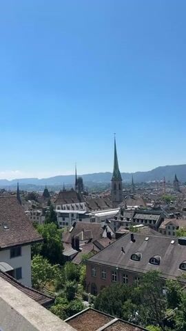The video shows an aerial view of a city skyline with a clear blue sky. The city has many buildings with distinct architectural styles, including churches with tall spires. Lush green trees are interspersed throughout the cityscape, suggesting a well-maintained urban environment. In the distance, rolling hills or mountains form a scenic backdrop. The camera pans slowly across the scene, offering a panoramic perspective of the city and its surroundings. There are no people or animals visible in the video. The overall impression is one of a peaceful and picturesque European city on a sunny day.