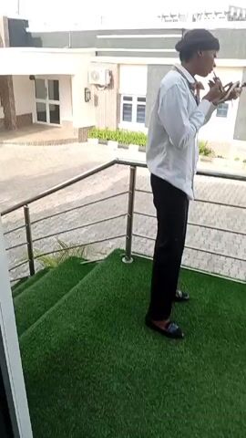 A woman films a man who is standing on an outdoor patio. He wears a light blue shirt, black pants, and a black head covering. He is holding and looking at a phone, while standing in front of a railing that surrounds the patio. Behind him are several buildings with white walls and air conditioning units. The camera is focused on the man, while the woman is slightly out of focus in the foreground.