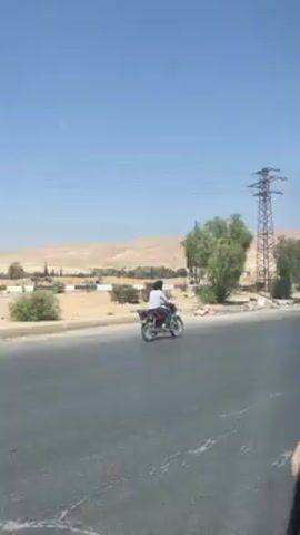 The video shows a person riding a motorcycle on a road. The motorcycle is dark-colored with silver accents, and the rider is wearing a white shirt and dark pants. The road is asphalt and has some cracks. There is a power line tower and trees next to the road. In the background, there is a desert landscape with hills. A blue road sign is visible, displaying the Arabic words for "Damascus" and "Quneitra" along with arrows pointing in opposite directions. The sky is blue. The video seems to have been taken from a moving vehicle.