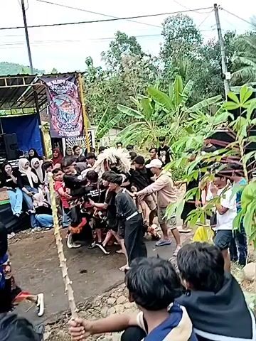 The video shows an outdoor gathering with a stage set up in the background. A crowd of people, mostly young adults and teenagers, are gathered around a central area where a performance is taking place. Two individuals are engaged in a traditional dance or performance, dressed in colorful costumes. The crowd appears to be watching intently, with some people recording the event with their phones. The environment is somewhat rural, with trees and houses visible in the background. The overall atmosphere seems energetic and festive.