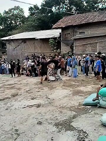A group of people are gathered in an outdoor area, watching a performance. In the center, two individuals are interacting with a person dressed in a costume that appears to be a horse. The costume is predominantly white with black and red striped detailing. The person in the costume is bent over and supported by the other two individuals. Several other people are standing and watching the performance, some are seated on the ground. The environment appears to be a village or rural setting with simple buildings in the background. The ground is dirt and uneven.