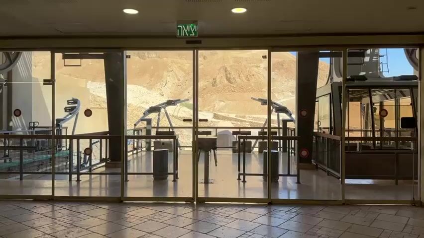 This video shows the interior of a cable car station with large glass doors opening to a mountainous desert landscape. Through the doors, a cable car can be seen to the right, and two sets of angled mechanical arms, likely part of the cable car system, are visible. There are railings and trash cans on the platform outside the doors. Inside the station, the floor appears to be tiled. Above the doors, an illuminated exit sign is visible. The overall scene suggests a transportation hub in a arid, rocky environment.