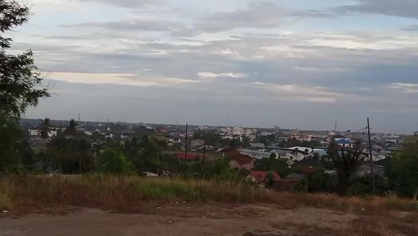 The video shows a panoramic view of a small city or town nestled among lush greenery, taken from a slightly elevated position. The cityscape includes various buildings, some of which are residential homes with red rooftops, and structures that appear to be commercial or public buildings distinguished by white domes. Numerous tall transmission towers are seen piercing the skyline, indicating the town’s connectivity. The foreground consists of dry, grassy terrain that gradually slopes down, revealing more greenery including trees and shrubs. A small body of water, possibly a pond or lake, is also visible, adding to the naturalistic environment. The sky above is filled with a layer of thin, overcast clouds, creating a soft, diffused light across the scene. The overall atmosphere is peaceful and somewhat serene.