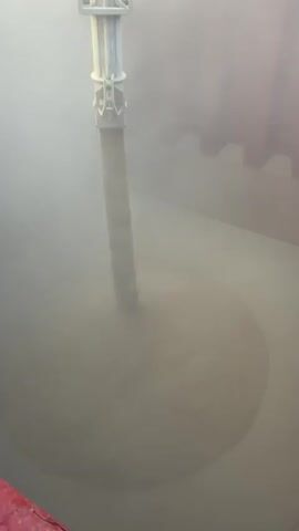 The video shows a bulk carrier ship being loaded with a light-colored powder, likely grain or a similar commodity. A large mechanical loading arm, a common piece of equipment used in port operations, is visible extending into the ship's hold and dispensing the material. The process creates a significant amount of dust, obscuring much of the interior of the hold and giving the scene a hazy appearance.

The external shots reveal the ship docked at a port, with other industrial structures and cranes in the background, suggesting a busy shipping terminal. The sky is visible, showing a clear blue with some wispy clouds, and the lighting indicates it's daytime, likely late afternoon given the warm hue of the light. The presence of silos in the distance further confirms the port setting and the large-scale nature of the operation.

No people are clearly visible within the ship's hold, though it's implied that dockworkers are operating the machinery from above or a control room. The overall mood is one of industrial activity and efficiency. The location is confirmed as Constanța, Romania, a major port on the Black Sea known for its bulk cargo handling. There is no discernible text in the video.