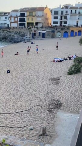 The scene is a sandy beach with a group of young men playing soccer. Several are actively engaged in running after the ball. One player is on his hands and knees. Another stands to the side, holding a phone. Further up the beach, several people are lying on towels. To the left, there is a rocky outcrop leading into the ocean. Behind the beach, there is a row of buildings of varying colors, mostly white and yellow. In the foreground, there is a backpack lying on the sand. The weather appears to be clear and sunny.