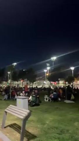 Here is a description of the video:

The video shows a large crowd of people gathering on a grassy area at night. Many people are seated on blankets or chairs, some at tables, seemingly enjoying a picnic or event. The area is well-lit by tall lampposts, and buildings can be seen in the background, including what appears to be a mosque's minaret. The atmosphere is relaxed and social, with individuals and groups chatting and eating. Overall, the scene suggests a community gathering or public celebration taking place in a park or recreational area after dark.