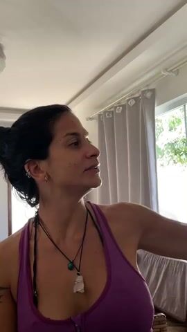 A woman with dark hair pulled back, wearing a purple ribbed tank top and multiple necklaces with pendants (one turquoise, one white/clear crystal), is featured in the video. She has earrings and visible freckles or sunspots on her face.

She holds a small, golden-brown fried fish in her right hand, bringing it up to her pursed lips with her eyes closed, as if playfully kissing or about to taste it. Her expression conveys a sense of amusement or anticipation.

The scene is indoors, brightly lit by natural light streaming through a window with white curtains in the background, suggesting it is daytime. The general setting appears to be a home. No visible text is present. The context provided indicates the location is Cabo Frio, Brazil.