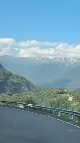 The video shows a view from inside a vehicle moving along a winding road through a mountainous landscape. The road is paved and has a metal guardrail on the right side. On the left side of the road, there are steep, tree-covered slopes, with some terraced areas visible. In the distance, a range of majestic, jagged mountains stretches across the horizon under a bright blue sky dotted with fluffy white clouds. As the vehicle continues, power lines are visible on the side of one of the mountains. A few buildings and houses are scattered throughout the valleys and on the hillsides. A black car is seen driving ahead on the road. The overall impression is one of a scenic drive through a natural, mountainous region on a clear day.