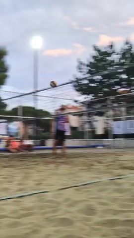 Two men are playing beach volleyball. One man, wearing a white tank top and black shorts, is in the foreground, preparing to hit the ball. Another man, wearing a white tank top and black shorts, is behind him. A volleyball is in the air above them. In the background, there is a volleyball net, trees, a light pole, and spectators. The game is being played on a sand court.