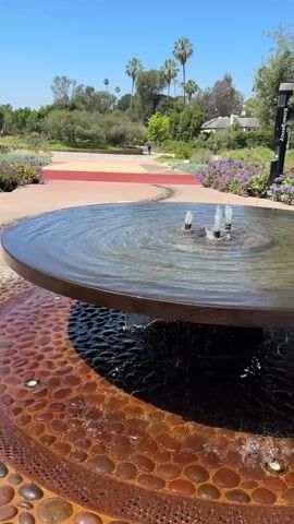 A circular fountain with multiple jets of water flowing upwards is in the foreground. The water cascades over the edge of the fountain and down into a lower basin adorned with decorative stones. The fountain is set in a park-like environment with lush green trees and a clear blue sky. In the background, a paved pathway winds through flower beds. A person is visible walking in the distance on the path.