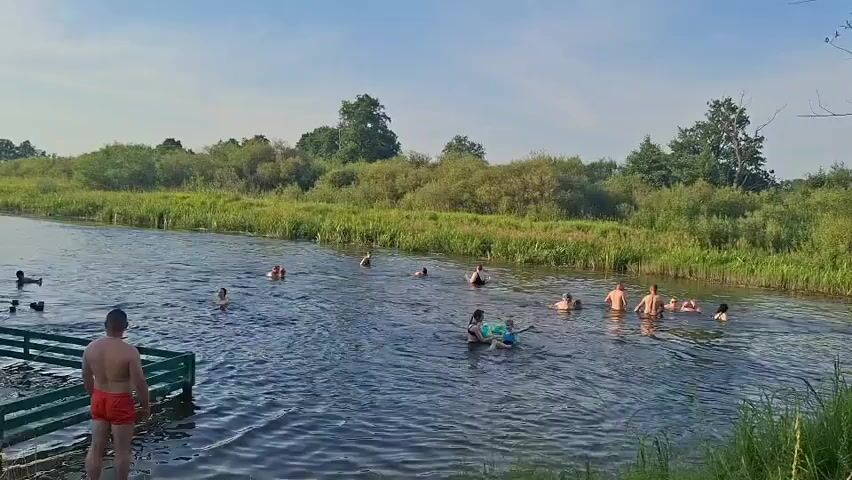A group of people are swimming and playing in a river on a sunny day. The river is surrounded by lush green vegetation and trees. In the foreground, a man in red swim trunks stands on the riverbank, facing away from the camera. In the water, several people are swimming, some with inflatable toys. The water is clear and appears to be shallow in some areas. There is a wooden structure, possibly a pier or a bathing platform, in the water on the left side of the image. The sky is blue with a few wispy clouds.