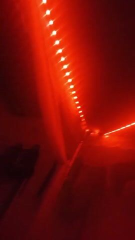 The video shows a room illuminated by LED strip lights that change color from green to red to blue. The lights are mounted on the ceiling and walls, casting a glow over the room. Visible in the shadows are clothing items and what may be furniture.  There is also speech audible.