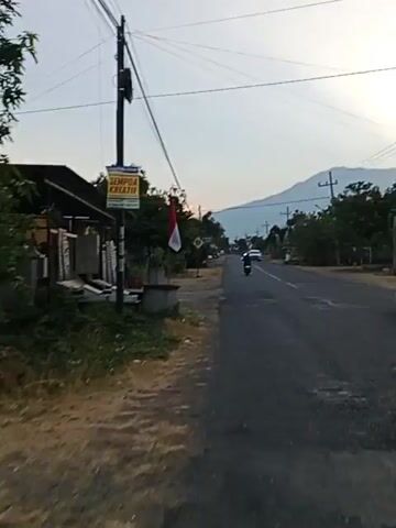 A video captures a street scene at what appears to be dusk or dawn. A paved road runs diagonally from the bottom right to the upper left of the frame. On the left side of the road, there is a dirt path and then a grassy area with some plants and a building with a dark roof and a yellow sign that reads SEMPOA KREATIF. A red and white Indonesian flag is hanging from a utility pole on the left. Further down the road, there are trees, streetlights, and other buildings. Several vehicles, including motorcycles and cars, are visible on the road, moving in both directions. In the distance, there are mountains under a light-colored sky. The camera appears to be mounted on a moving vehicle, likely a motorcycle, as indicated by a blurred object in the bottom right corner of the screen that resembles part of a helmet or vehicle. A traffic sign with a silhouette of two people walking is visible on the left side of the road.
