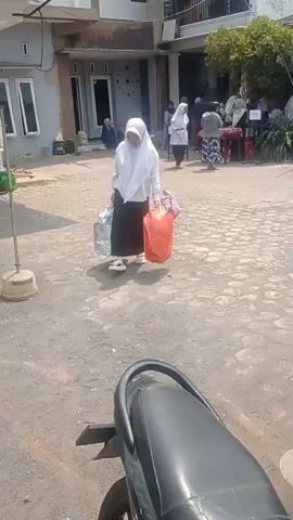 A woman wearing a white hijab and black pants walks across a paved outdoor area, carrying two shopping bags and an orange plastic bag filled with items. She is walking away from the camera. In the background, a group of people are standing or sitting near a building. Some individuals are wearing hijabs. There are tables and chairs visible in the background. The ground is a mix of pavement and dirt. A portion of a motorcycle is in the foreground. The lighting suggests it is daytime.