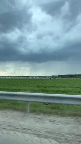 The video shows a drive on a highway. The weather is overcast and stormy. Fields of green crops are visible through the car window. Other vehicles are also on the road ahead. The camera focuses on the road and the view from the front of the car.