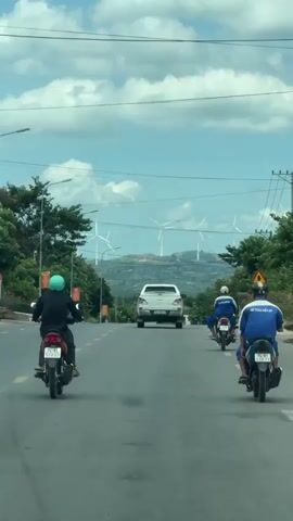 The media captures a daytime scene on a paved road in Lao Bảo Commune, Vietnam. Several individuals are riding motorcycles, heading in the same direction down the road. In the foreground, three motorcycles are prominent, with riders wearing helmets and casual clothing. One rider in the center is wearing a black shirt and a green helmet, while the two riders on the right are in blue shirts. A white pickup truck is visible further down the road, with a white van also present. In the background, a sprawling landscape unfolds with rolling hills dotted with numerous wind turbines, suggesting a renewable energy site. Lush green trees line the sides of the road, and power lines crisscross the sky, which is filled with fluffy white clouds under a bright blue sky. The overall impression is one of everyday movement and activity within a scenic, potentially rural or semi-rural Vietnamese setting. One of the blue shirts has visible white text, "SE TONG KIEN MY". License plates on the motorcycles are also visible, with one reading "74-H1 135.77". The lighting suggests it is daytime, likely late morning or early afternoon.