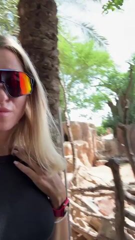 A woman with blonde hair and sunglasses is taking a selfie in front of a zoo enclosure. She is wearing a black top and a pink smartwatch. The enclosure appears to be for giraffes, with palm trees, grassy areas, and a small body of water visible. In the background, two giraffes can be seen. The scene is outdoors during the daytime, with lush greenery and sunlight.