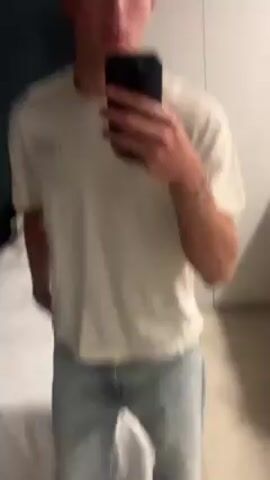 A young man takes a selfie video in what appears to be a bedroom. He is wearing a light-colored t-shirt and light wash jeans. A white wardrobe is behind him and a bed is to his left. The walls are painted a teal or dark blue color. The man runs his hand through his hair and moves the phone around to show more of his outfit.