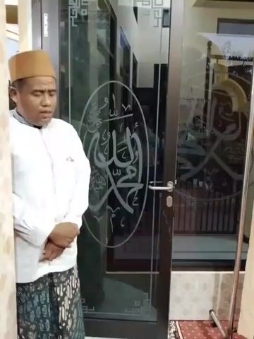 The video shows a group of men standing in what appears to be a prayer room or a religious gathering space. In the foreground, a man wearing a white shirt and a peci hat is standing with his eyes closed and hands clasped in front of him, seemingly in prayer. Behind him, a glass door with Arabic calligraphy is visible. To the right, a rack with beige curtains separates the area. Further into the frame, three men are standing in a line, also wearing peci hats and traditional clothing. They appear to be listening or waiting. The room has red carpet flooring with a patterned design. The lighting suggests an indoor setting. The video is in landscape orientation and the footage is somewhat shaky.