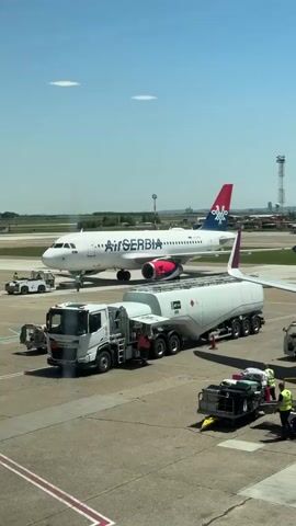 This media file captures an airport tarmac scene on a bright, sunny day. The main subjects are a white Air Serbia airplane, a white fuel truck, and several individuals in high-visibility vests, likely airport ground crew. The airplane, with "Air Serbia" prominently displayed on its fuselage and a red and blue tail fin, is parked on the tarmac. Adjacent to the plane is a large, white fuel truck with a long cylindrical tank, indicating refueling operations are underway or about to commence.

Several people in yellow vests are visible around the aircraft and the fuel truck, engaged in various tasks associated with aircraft servicing. One person appears to be connecting or disconnecting equipment from the fuel truck. Another individual is standing near a baggage cart. The overall scene suggests a routine operational moment at an airport, preparing an aircraft for its next flight.

The setting is clearly an airport tarmac, with paved surfaces marked with yellow and white lines. In the background, other airport structures, including a control tower and distant buildings, are visible, along with other vehicles and aircraft. The clear blue sky and bright sunlight indicate it is daytime, likely midday. The weather appears to be fair and warm.

Notable details include the clear visibility of the Air Serbia branding on the aircraft. The fuel truck also has some visible text and markings, though not entirely legible at this resolution. The presence of ground crew in safety vests emphasizes the operational nature of the scene. There are no explicit emotional cues conveyed, but the activity suggests efficiency and preparedness. The location context provided points to Surcin Urban Municipality, Serbia, which would be consistent with an airport like Belgrade Nikola Tesla Airport.