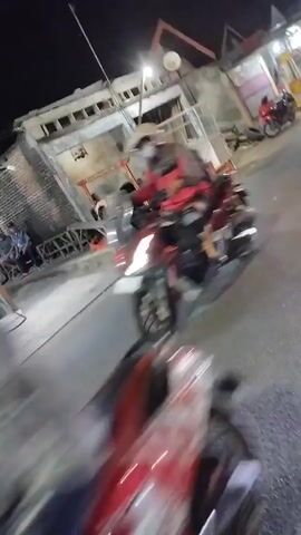 The video appears to be a short, shaky, first-person recording taken at night from a moving vehicle, likely a motorcycle, on a street with buildings on the right. The focus is blurry due to motion, but several motorcycles and a silver car are visible. On the right side of the street, there is a building with an open front, possibly a shop or food stall, with people seated inside and around it. One motorcycle passes the recording vehicle, carrying two people. Another motorcycle, also carrying two people, is seen further down the street. A silver car is also observed moving along the street. The overall scene suggests a lively evening setting with people out and about.