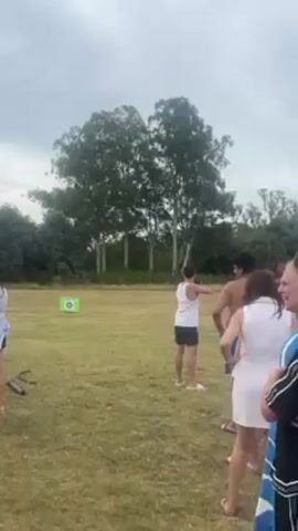 The video shows a group of people standing on a grassy field near some trees. One man is standing in front of them, aiming an arrow at a target in the distance. The other people are watching him. The sky is overcast.