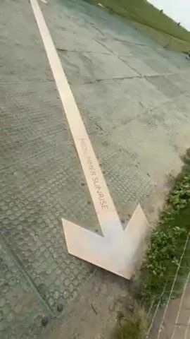 The video shows an arrow on the ground pointing towards Stonehenge in England. The arrow is beige with the words "MIDSUMMER SUNRISE" written on it, and it's placed on a green plastic grid. In the distance, the iconic Stonehenge monument is visible on a grassy field under a cloudy sky. There are also a number of people milling around, and it seems that this arrow might be pointing to where the sun rises on Midsummer.