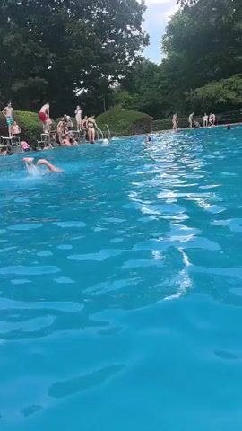 The video shows a busy outdoor swimming pool on a sunny day. The camera is positioned low, just above the water's surface, giving a dynamic perspective of the pool. The water is a vibrant blue, with ripples and reflections suggesting movement. In the background, several people are visible swimming, standing, and interacting in and around the pool. A diving board with a platform is prominent on the right side of the frame, with a few individuals gathered at the top. Further back, a concession stand or small building with a red awning is visible, with more people nearby. Lush green trees form the backdrop, indicating a natural, park-like setting. The overall atmosphere appears lively and recreational, with many people enjoying a summer day at the pool.