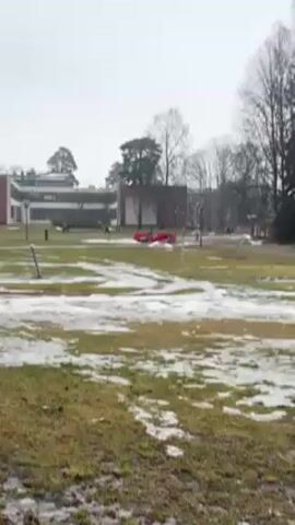 The video shows a park or large lawn on an overcast day with patches of melting snow scattered around. There are brick buildings in the background. The camera pans to the right, showing a muddy path marked by small orange poles.  The path leads towards a wooded area with tall bare trees. The ground is still covered with patches of snow and standing water.