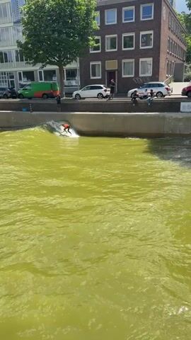 The media file shows a person surfing on a stationary wave in a canal. The surfer, wearing an orange top and black bottoms, is actively engaged in riding the wave, exhibiting a crouched stance and outstretched arms for balance. The water is a vibrant, almost neon green, suggesting it may be treated or artificially colored, or perhaps reflecting the surrounding foliage.

The scene is set in an urban environment, likely Rotterdam, Netherlands, as indicated by the location context. In the background, a multi-story building with numerous windows and a distinct architectural style stands against a clear, sunny sky, implying daytime. Several cars are parked along the embankment, and other spectators are visible on the walkway above the canal, observing the surfing activity. The presence of people on the walkway and cars suggests a public area, possibly a park or recreational space within the city.

Notable details include the bright, sunny weather, which contributes to the overall lively atmosphere. The water's unusual color is a striking feature. Various signs are visible on the embankment, with legible text including "DUX GROUP," "THE BASTARD," "ZAPPLY," "BREITLING 1884," and "PORSCHE Centrum Rotterdam." These signs indicate sponsorships or businesses associated with the location or event. The overall mood appears to be one of active recreation and enjoyment on a pleasant day.