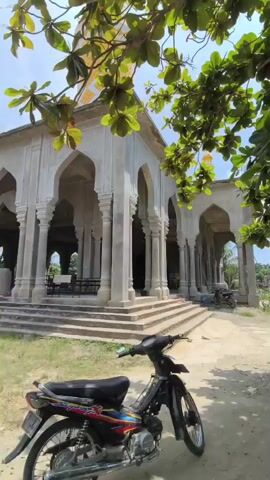 The video shows a partially constructed mosque with a minaret. In the foreground, a motorcycle is parked on a dirt path. Lush green foliage partially obscures the view of the mosque from the top. The mosque has a series of arched openings supported by columns and a dome. The minaret is tall and yellow. The sky is bright and the weather appears to be sunny.