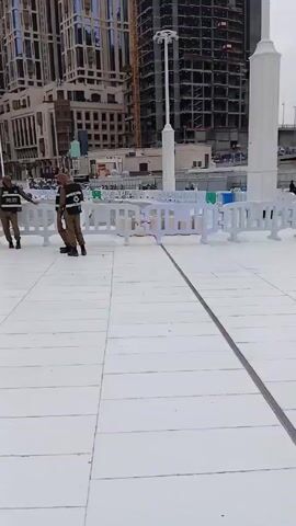 The video shows a plaza in front of a large building. The plaza is paved with white tiles. The building has a gray stone facade with large arched windows. There are tall white pillars with lights on top scattered throughout the plaza. The video shows people walking around, some in traditional Islamic clothing. There are also security personnel present. White and green plastic barriers are used to cordon off certain areas. The sky is overcast.