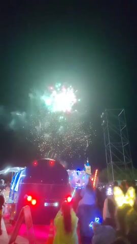 The video captures a nighttime outdoor event with fireworks illuminating the dark sky. In the foreground, a crowd of people are visible, some holding balloons and some wearing mouse ears. In the middle ground, a large, dark vehicle, possibly a truck, is parked, with its red taillights illuminated. Above the vehicle and the crowd, fireworks explode in a dazzling display of pink, white, and green lights, with trails of smoke ascending into the night. In the background, a tall, metallic structure resembling a stage or lighting rig stands prominently. Further in the distance, a building with a blue dome and spire is visible, suggesting a potentially festive or event-oriented location. The video appears to be shot from a low angle, looking up at the fireworks.