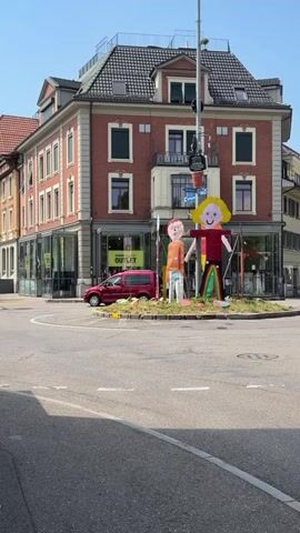 The video shows a roundabout with a large statue of two figures, one adult and one child, in the center. The adult figure is taller and has a yellow head with a smiling face, a red and black torso, and a colorful striped skirt. The child figure is smaller, with a round head and an orange and brown outfit. Both figures appear to be made of lightweight, colorful materials. Behind the roundabout, there is a multi-story brick building with shops on the ground floor and apartments above. A red van is parked on the side of the road. The sky is clear and blue, indicating a sunny day. Traffic cones and debris are scattered around the base of the statues, suggesting recent construction or maintenance. Cars are seen driving around the roundabout and on the streets. The overall scene depicts an urban environment with a touch of whimsical public art.
