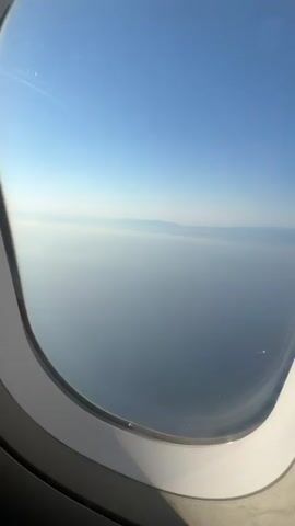 The video shows a view from an airplane window looking out at the sky and a body of water. The sky is clear and blue, with some wispy clouds near the horizon. The water is a deep blue and appears calm. There are faint outlines of land visible in the distance, possibly mountains. The bottom of the frame shows the edge of the airplane window. The video appears to be taken during the daytime, likely in the morning or afternoon.