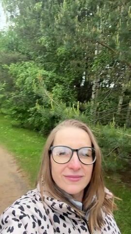 A woman films herself walking on a trail in a park. The woman has brown hair and is wearing black glasses and a black and white spotted jacket. The trail is dirt and lined with trees. There is green grass and a body of water is visible in the background. A man walks in the background.