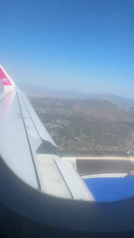 The video shows an aerial view from the window of an airplane. The clear blue sky fills the upper part of the frame. Below, the wing of the plane is visible, marked with "SKY" in pink lettering on the winglet. The wing is white and shows some signs of wear. Part of the engine is also visible beneath the wing, featuring blue accents. The landscape below is a mix of urban and rural areas. Densely packed buildings contrast with open fields and mountains in the distance, all covered with a hazy veil.