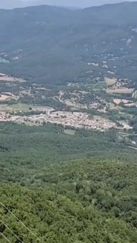 An aerial view shows a dense forest covering rolling hills. In the distance, a small town is visible nestled in a valley, surrounded by fields. The sky is hazy and overcast. The camera appears to be moving, panning slightly across the landscape. Power lines are visible in the foreground, suggesting the camera is on a vehicle or structure with a vantage point.