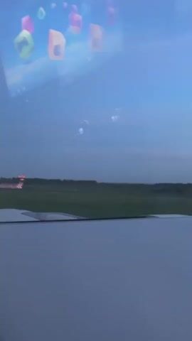 The view from inside a plane, looking out the window onto an airfield. The plane is approaching the runway for landing. Outside, the sky is blue with a hint of twilight. The wing of the plane is visible in the foreground. Reflected in the window is a screen displaying various app icons.