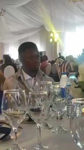 The camera pans across a table setting in a brightly lit, tented space, presumably for a wedding or formal event. A young man with short hair and a checkered jacket sits in the background. The table is elaborately set with numerous clear wine glasses, plates, water bottles, a menu with an animal logo, and a large floral arrangement of white flowers in a vase. Other guests are visible in the background, some in suits and dresses, sitting at other tables. The tent's ceiling is draped with white fabric, and several lights are visible.