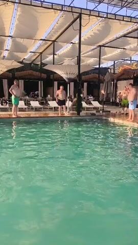 The video displays a pool area at what appears to be a resort or a public pool. The water in the pool is a clear turquoise color. Several individuals are visible. On the left side of the pool, a man in green shorts stands near the edge. In the center, two men in dark shorts are present, one of whom is interacting with a small palm tree. Further to the right, another man in blue shorts is standing, and to his right, a person with a pink object, possibly a child's toy, is visible.

The scene is set under a canopy supported by metal beams, with white fabric coverings providing shade. In the background, lounge chairs and cabanas are set up, suggesting a place for relaxation and leisure. The lighting indicates it is daytime, with sunlight filtering through the canopy and reflecting off the water. The overall atmosphere appears to be casual and relaxed, consistent with a poolside setting. The location is specified as Vahdat, Tajikistan. There is no visible text in the video.