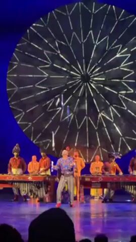 A performance is happening on a stage. At least 9 people are visible. Most of them are in a line behind what appears to be xylophones. They wear colorful, vaguely ceremonial outfits and tall hats. They are playing the xylophones. Two people are in front of the line, interacting with each other in what appears to be a dance or performance. They move around each other while facing the audience. There is a large, round, decorative object behind the performers. The background is dark blue. The lighting is bright. There is an audience in the foreground.