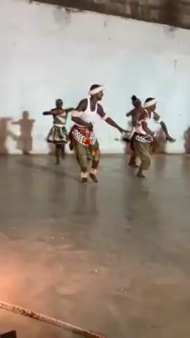 The video shows a group of six people performing what appears to be a traditional African dance in a large, bare room.  The dancers, a mix of males and females, are dressed in similar traditional attire that includes light-colored tops, patterned waist coverings or skirts, and headbands.  The floor of the room is a light, reflective concrete.  The room has white walls and a high ceiling.  A rope is visible in the foreground. The dancers move in synchrony, performing coordinated arm and leg movements.  Shadows of the dancers are cast on the back wall.